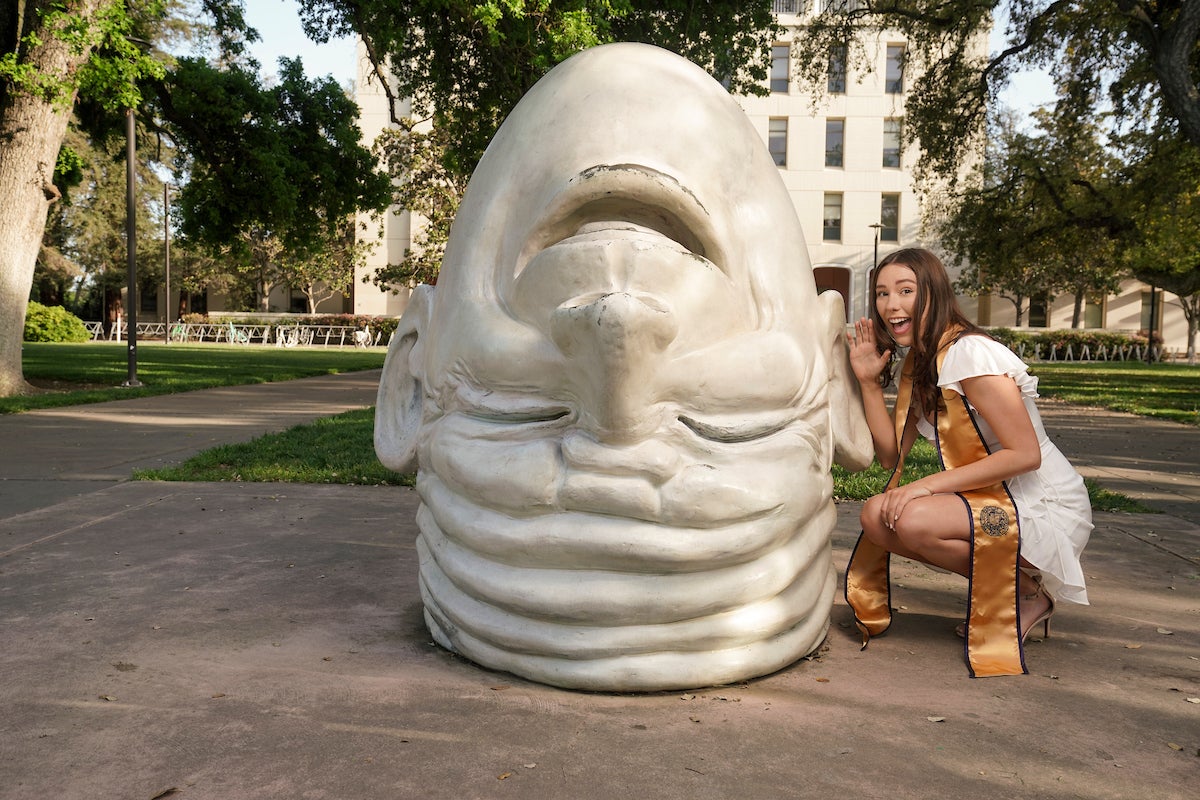 UC Davis graduating student Alexa Carter in white dress and graduation sashpretends to speak in Egghead sculpture's ear for a playful graduation photo